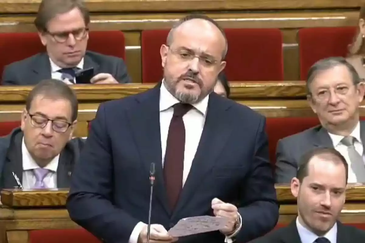 Un home de vestit i corbata parla en un parlament mentre altres persones ho observen des dels seients.