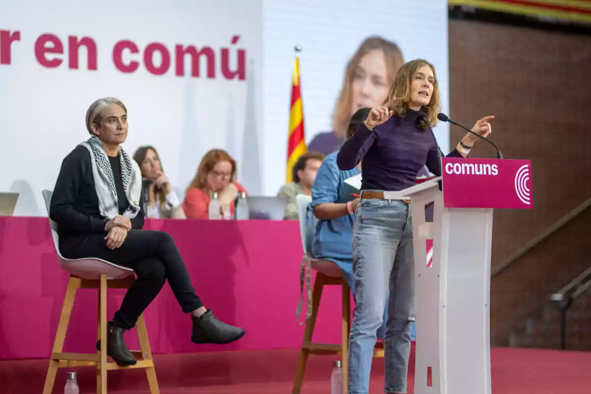 Jéssica Albiach parlant en un podi amb el logo de "comuns" mentre Ada Colau està asseguda a prop i diverses persones estan al fons.