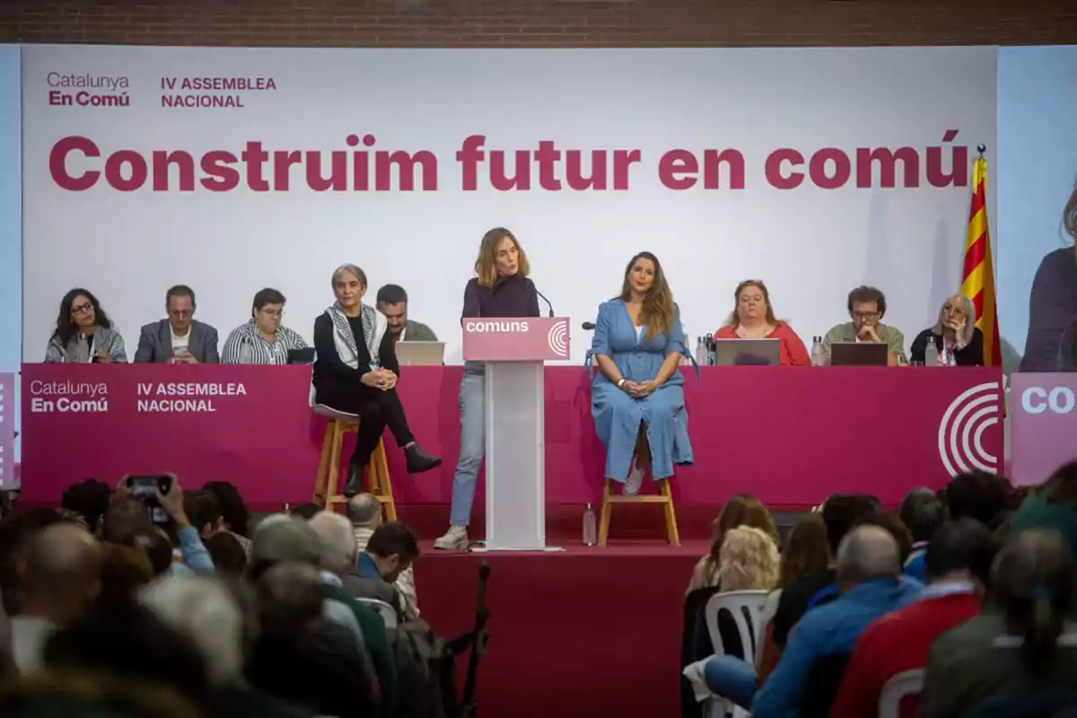 Una dona parla en un podi durant una assemblea de Catalunya En Comú amb diverses persones assegudes darrere seu i una audiència al capdavant.