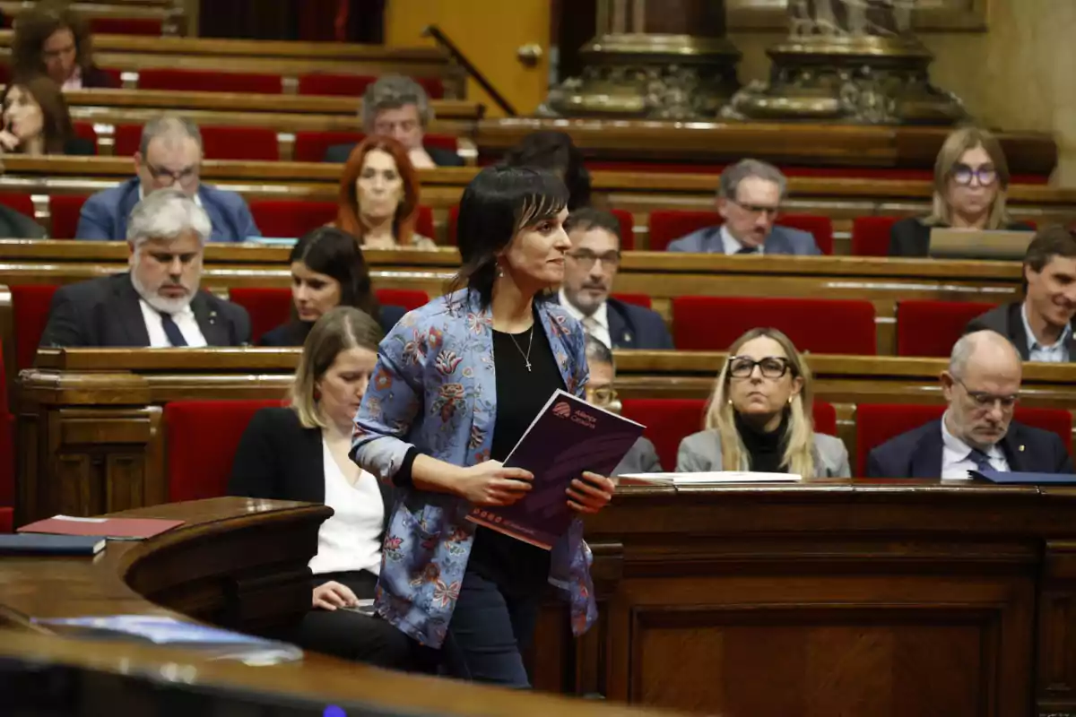 Una dona dreta sostenint documents en una sala de reunions amb diverses persones assegudes en banques vermelles.