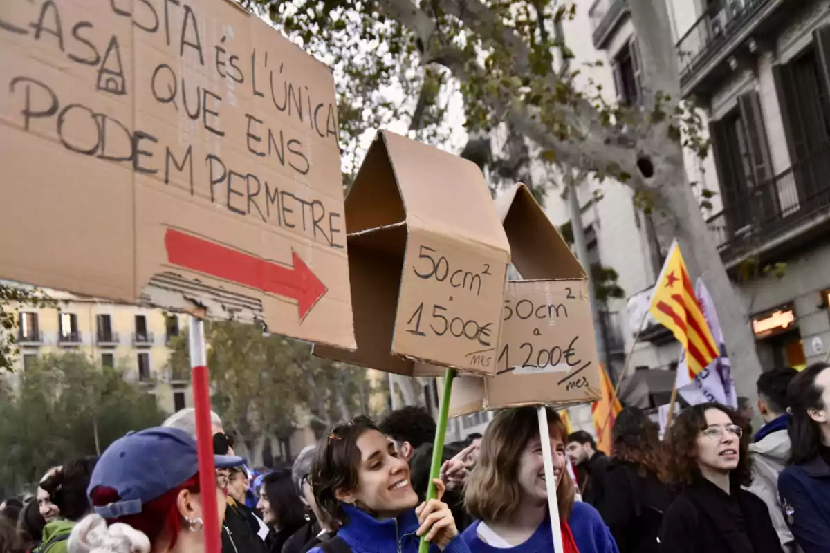 Persones en una manifestació sostenen cartells de cartró amb forma de casa amb missatges sobre l'alt cost de l'habitatge.