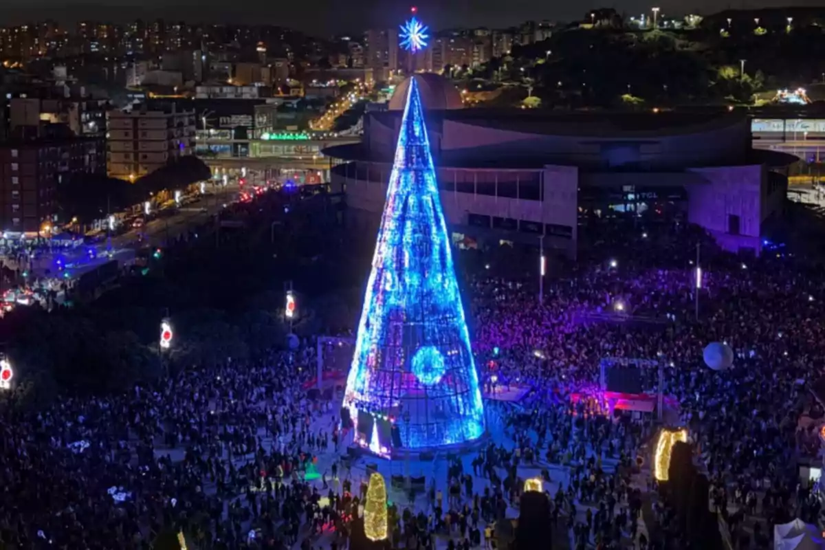 Una multitud es reuneix al voltant d'un gran arbre de Nadal il·luminat a una plaça durant la nit amb una ciutat de fons.