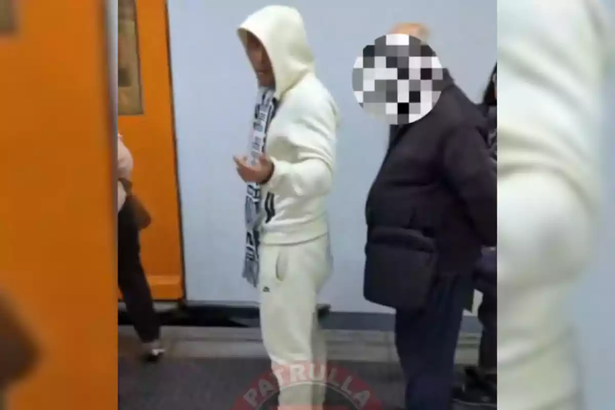 Dues persones dempeus en una estació de metro una amb dessuadora blanca i caputxa i l'altra amb el rostre pixelat.