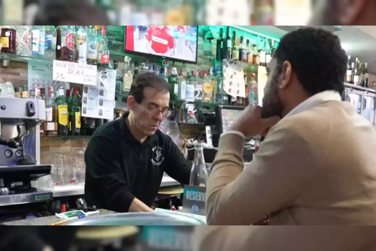 Un home darrere de la barra d'un bar atén un client mentre prepara una beguda, envoltat d'ampolles i una pantalla de televisió al fons.