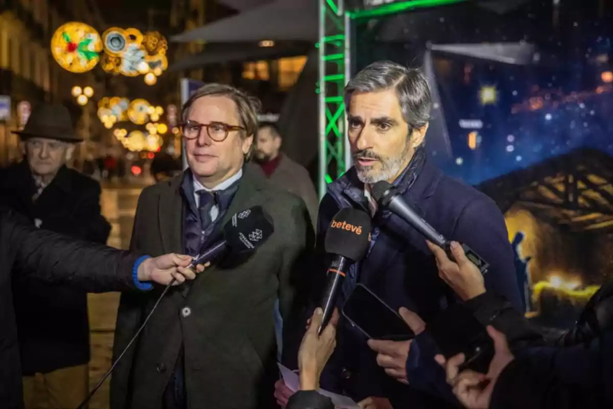 Dos hombres son entrevistados por varios medios de comunicación en un evento nocturno con luces decorativas de fondo.