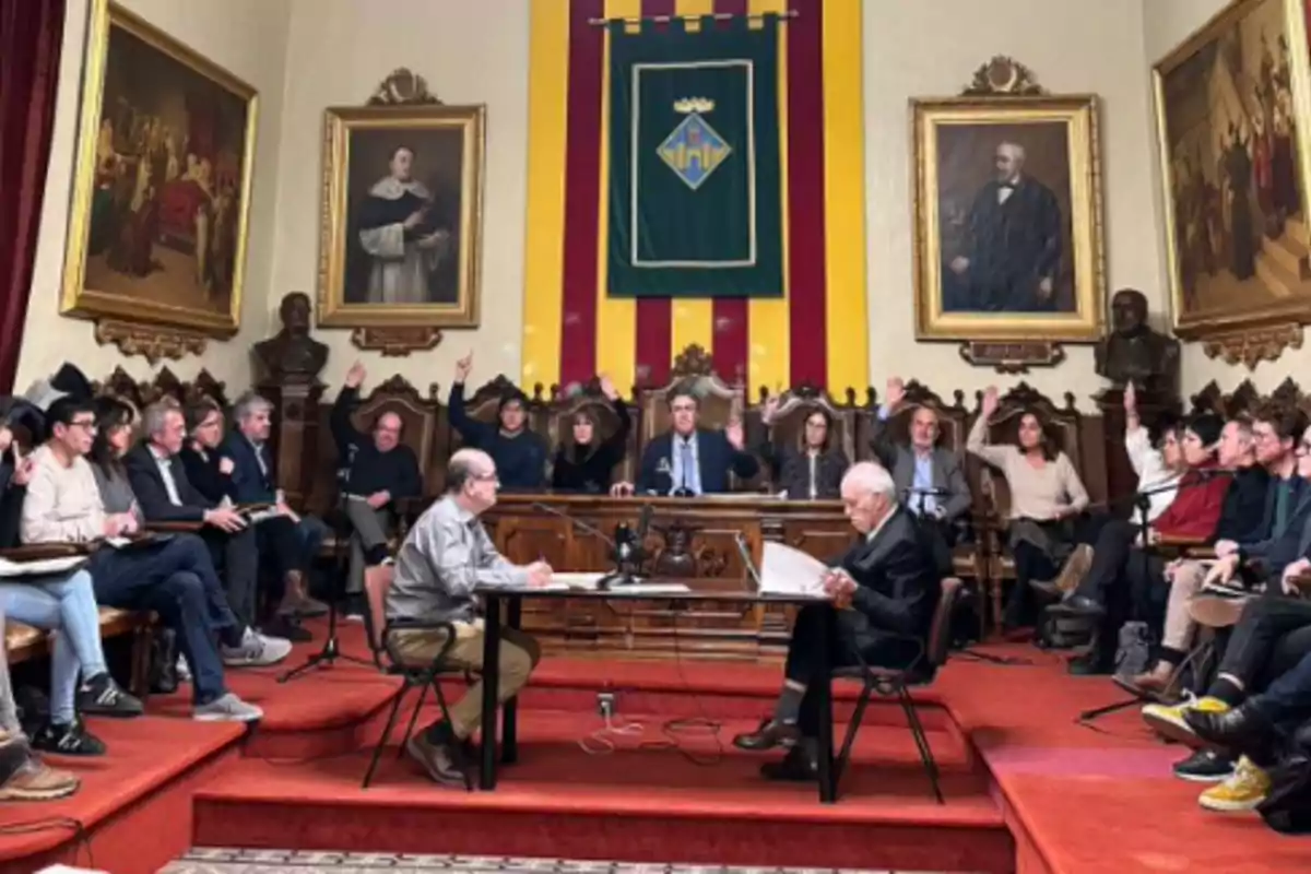 Un grup de persones en una sala de reunions amb retrats a les parets i una bandera darrere una taula central.