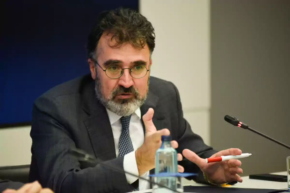 Un hombre con barba y gafas habla gesticulando en una reunión, con un micrófono y una botella de agua frente a él.