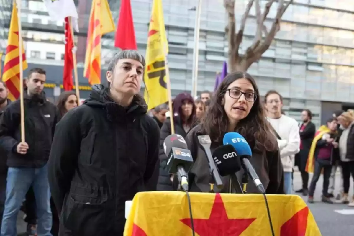 A group of people is gathered outdoors, some holding flags with yellow and red designs, while two women speak in front of several microphones.