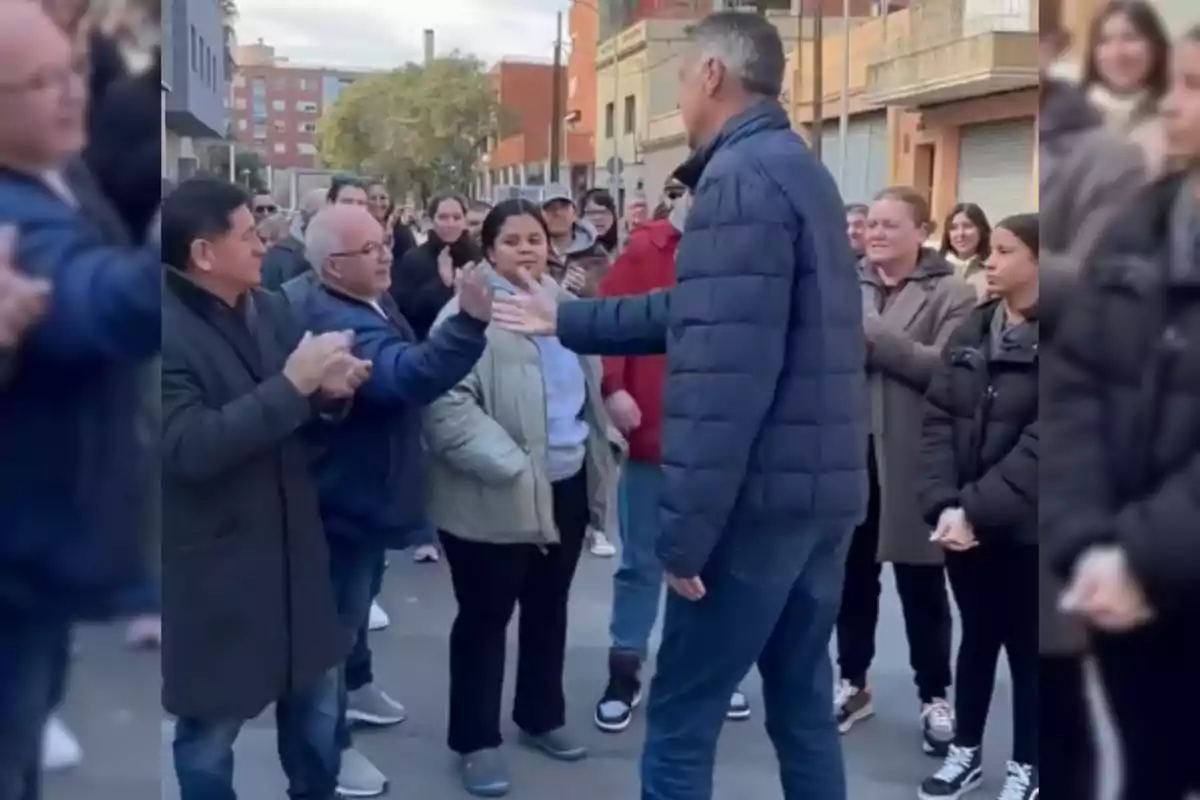 A group of people gathers in the street, some clapping while a tall man in a blue jacket interacts with them.