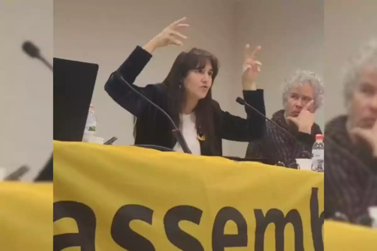 A woman gestures with her hands while speaking at a table with a microphone and a yellow sign, joined by another person who watches her.