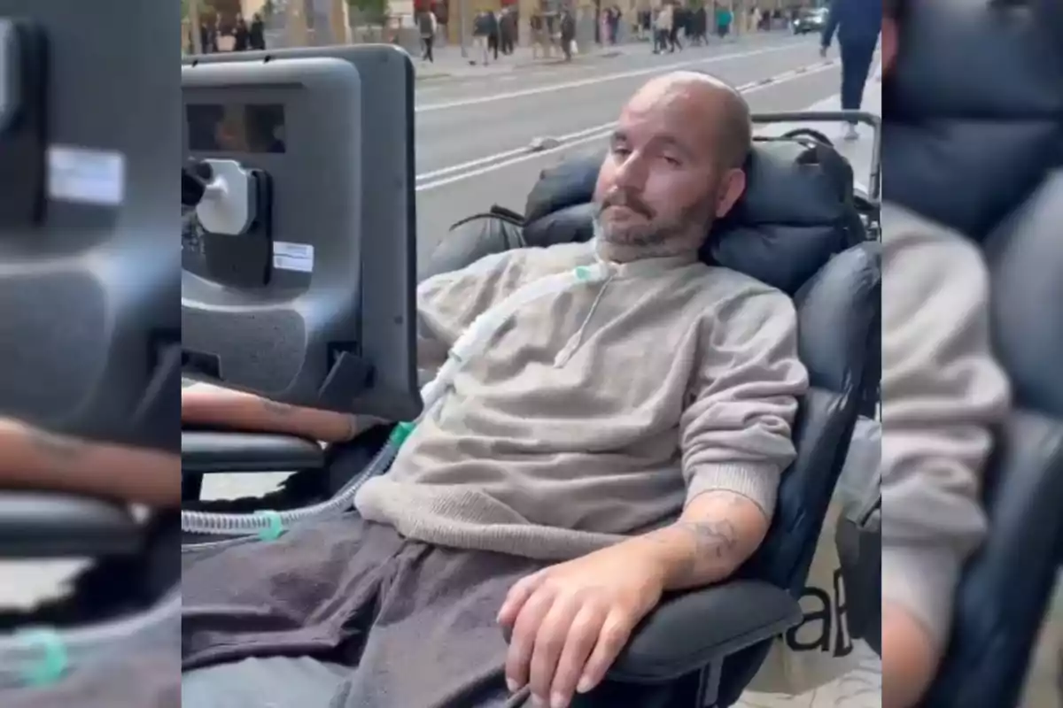 A man is reclining in a motorized wheelchair with a monitor in front of him and a breathing tube connected, while he is on a busy street.