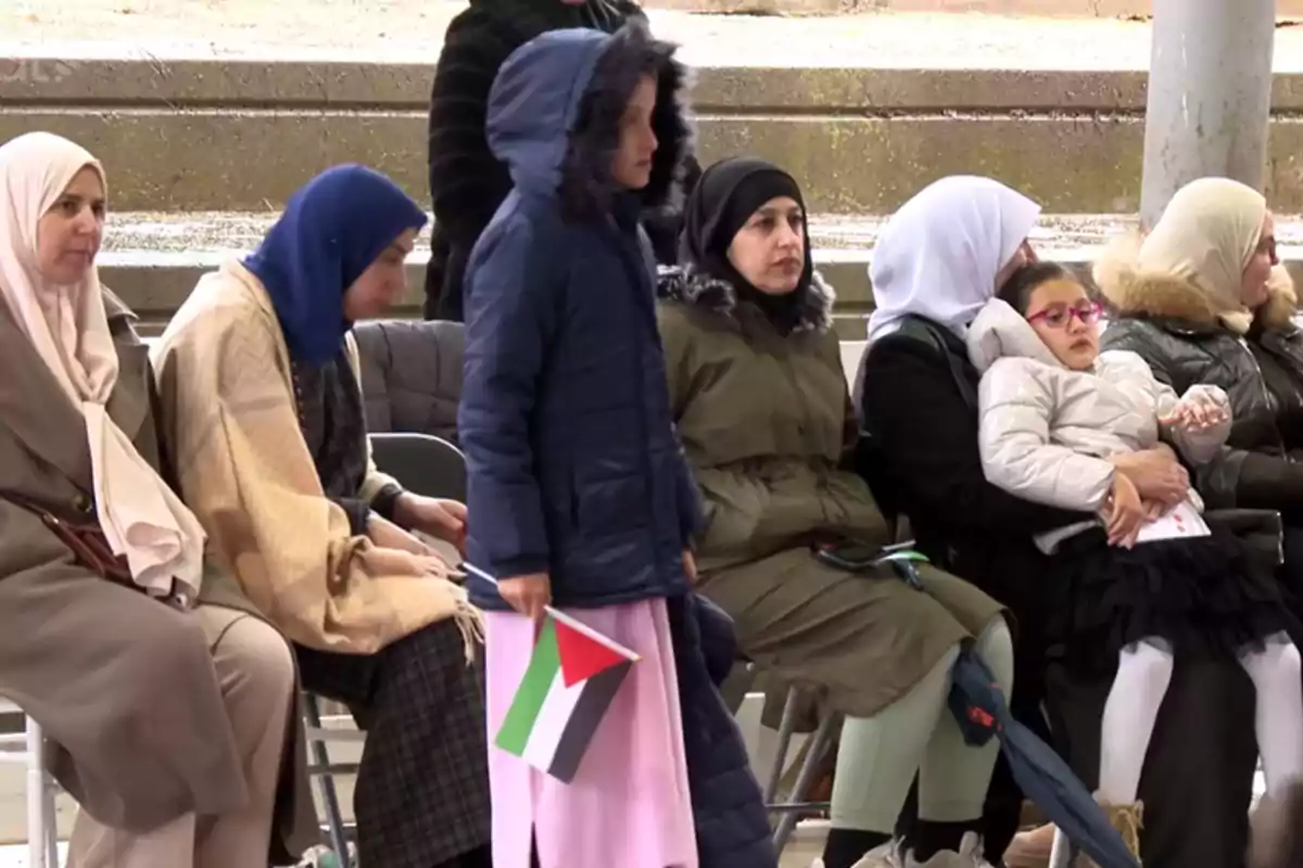 Un grupo de personas, algunas con hijabs, están sentadas y una niña de pie sostiene una bandera.