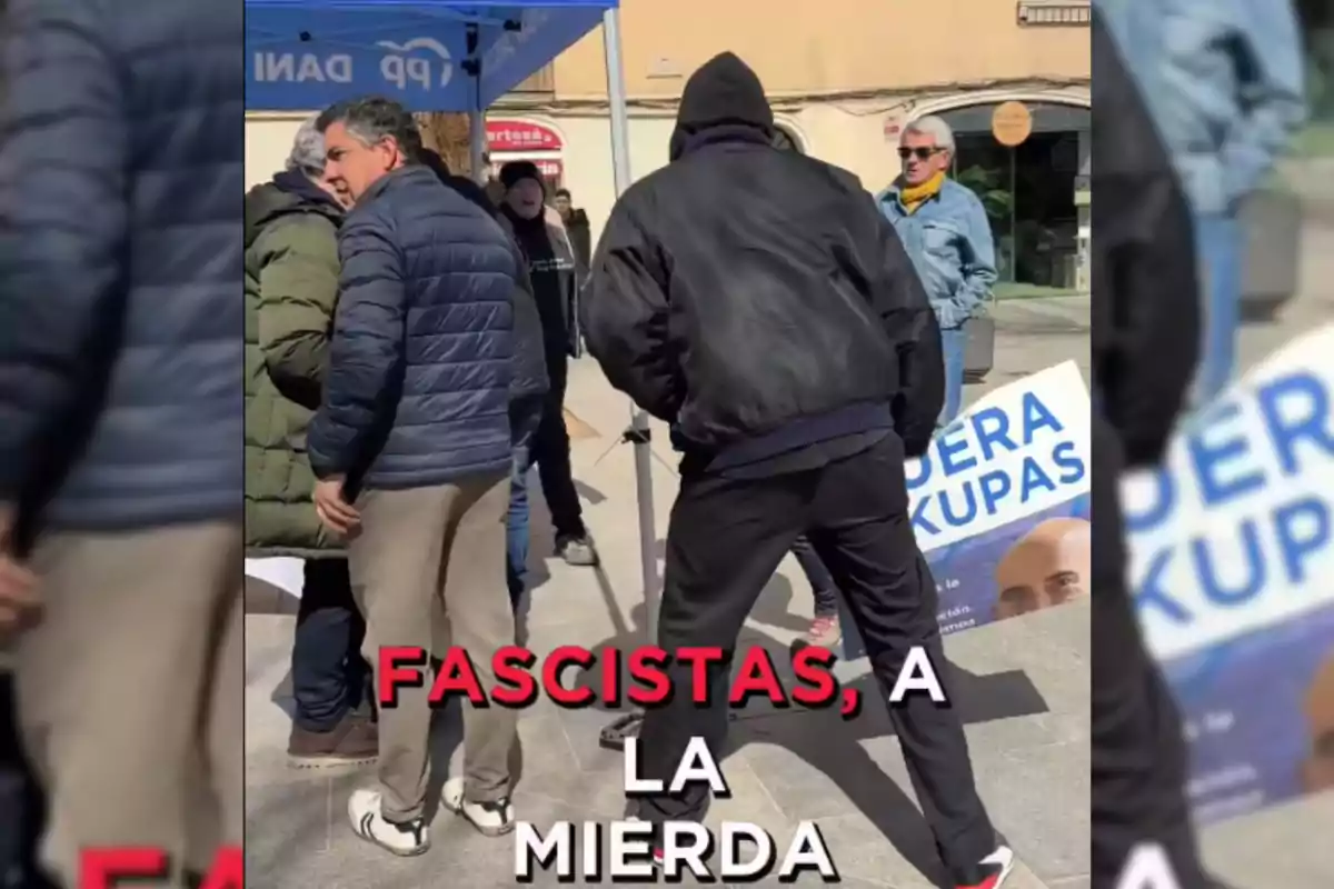 Un grupo de personas está reunido en una calle, algunas de ellas bajo una carpa azul con letras blancas, mientras que en el primer plano se ve a una persona con chaqueta negra y capucha; en el suelo hay un cartel parcialmente visible con texto en letras grandes.