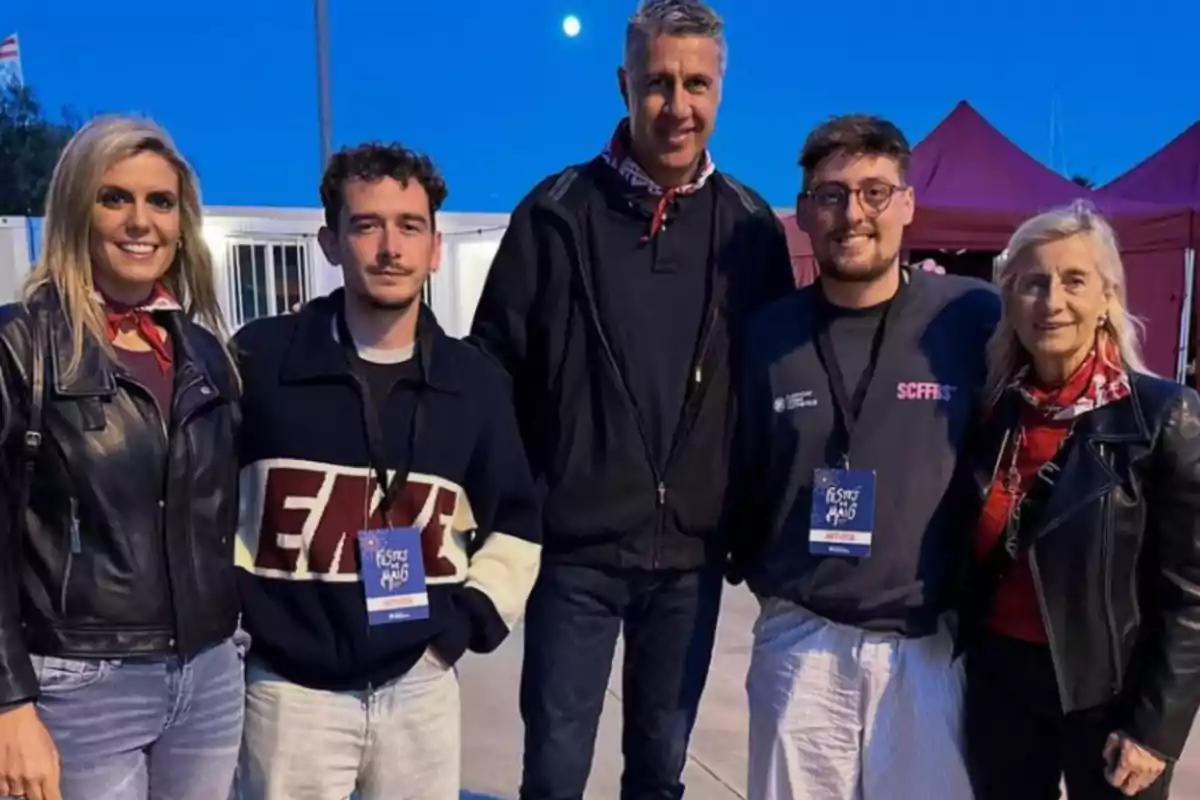 Un grup de cinc persones posa per a una foto a l'aire lliure durant un esdeveniment, amb un cel blau de fons i tendes vermelles darrere.