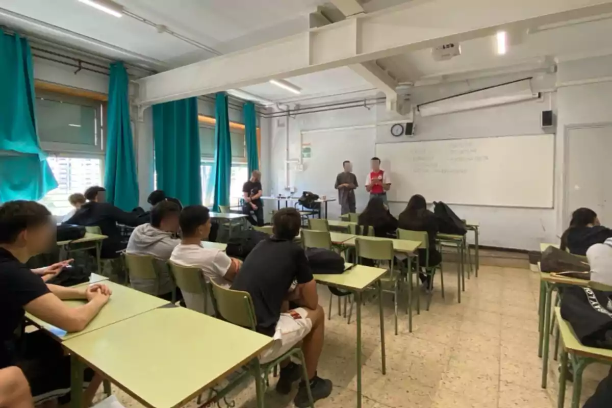 Un grup d'estudiants està assegut en una aula mentre dues persones presenten davant la pissarra i la resta observa des dels seus pupitres.