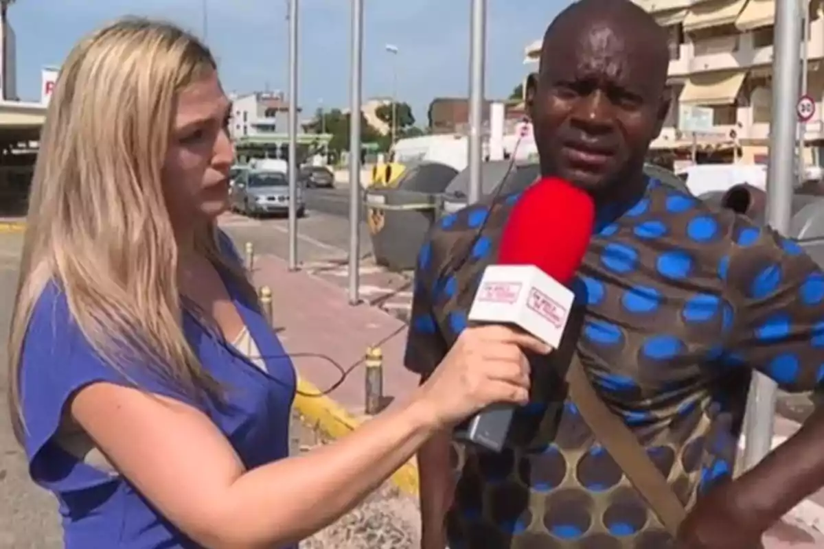 Una reportera sosté un micròfon vermell mentre entrevista un home al carrer durant el dia.