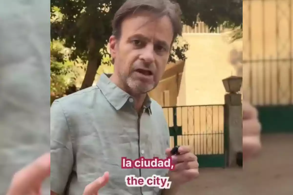 Brown-haired man with a short beard wearing a light gray jersey speaking outdoors in front of a green fence and trees, with Spanish and English subtitles that say "la ciudad the city"