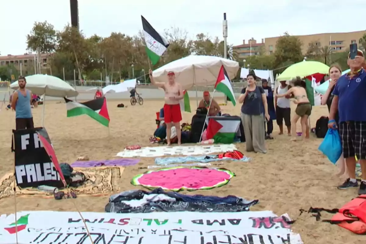 Persones en una platja amb banderes palestines i pancartes, algunes de les quals estan assegudes sota para-sols mentre d'altres estan dretes, una sostenint una bandera.