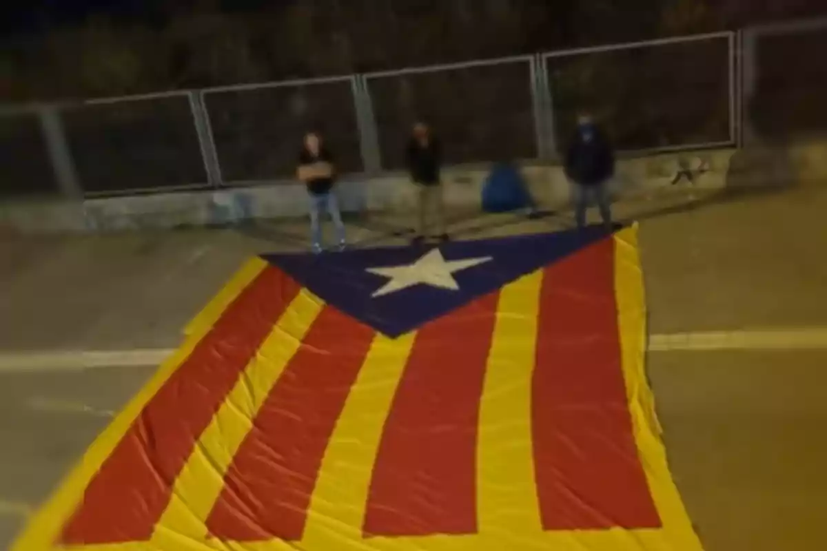 Tres persones dretes darrere d'una gran bandera estelada a terra, amb una tanca metàl·lica al fons.