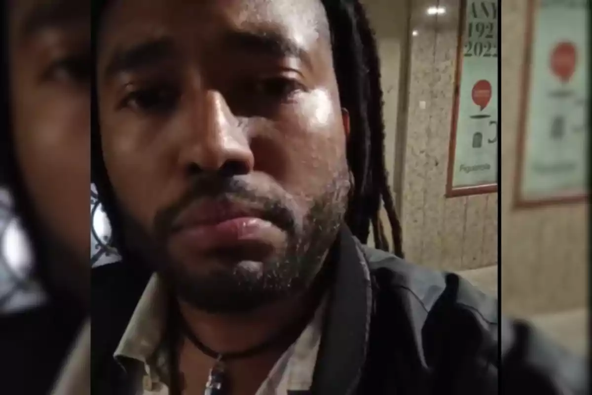Un hombre con rastas y barba mira a la cámara con una expresión seria, mientras está en un lugar con paredes de mármol y un cartel en el fondo.