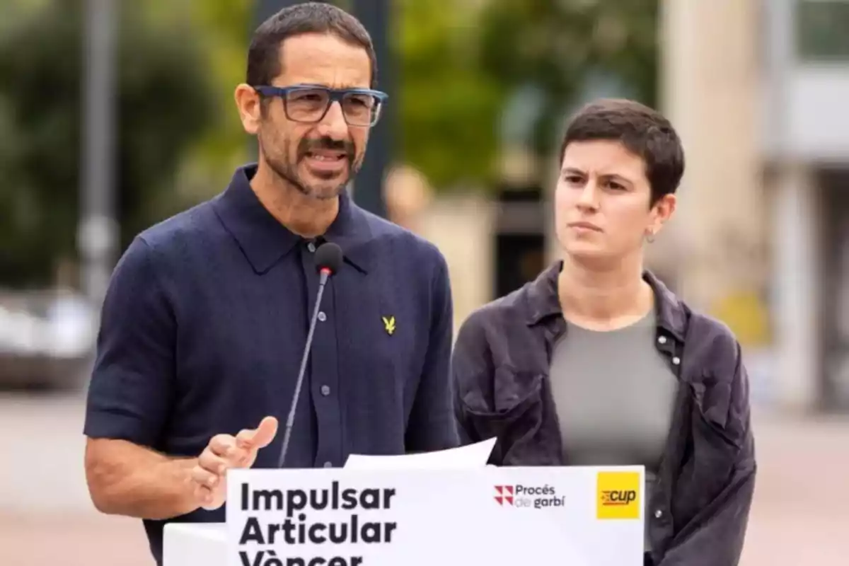 Dues persones en una conferència de premsa a l'aire lliure amb un micròfon i un cartell que diu “Impulsar Articular Vèncer”.