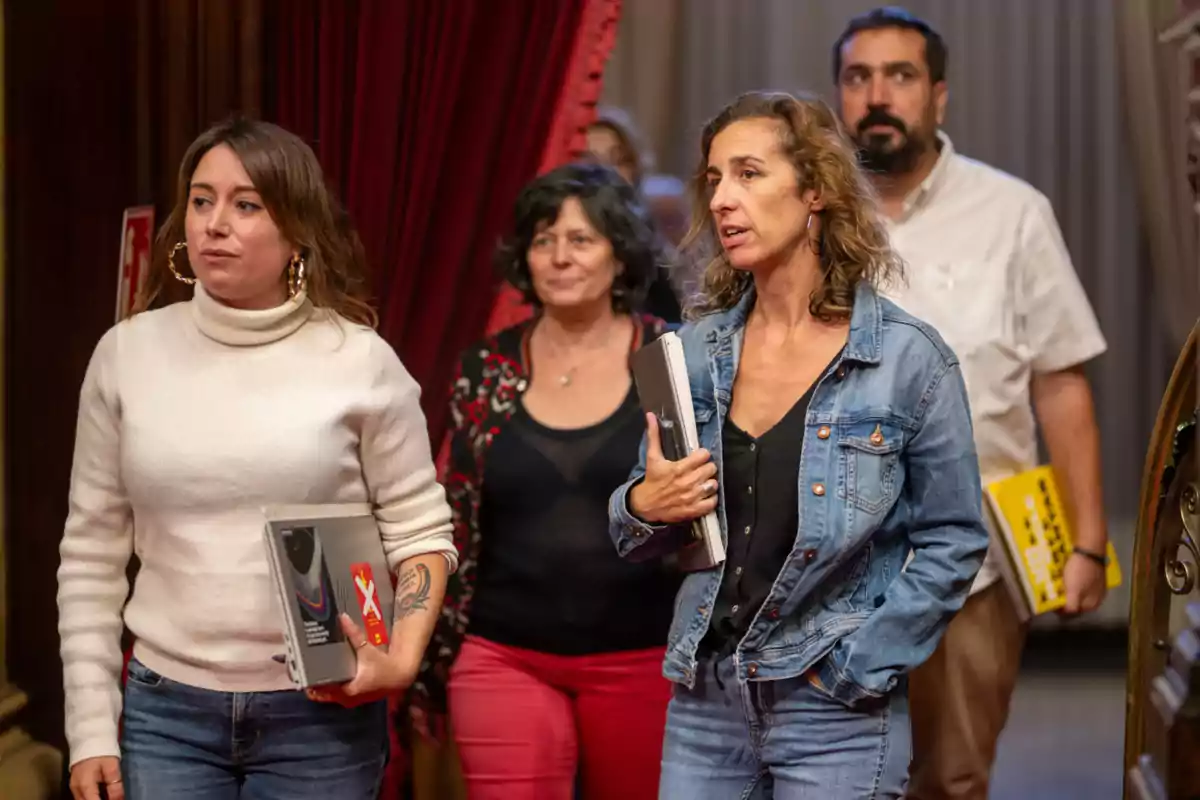 Un grup de persones camina per un passadís amb cortines vermelles, portant carpetes i llibres.