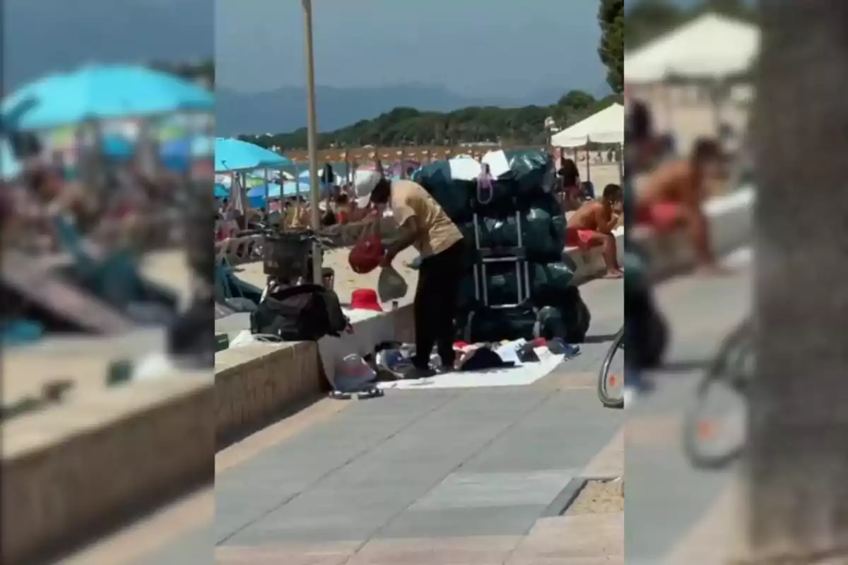 Una persona organitzant mercaderia en una platja concorreguda amb para-sols i turistes al fons.