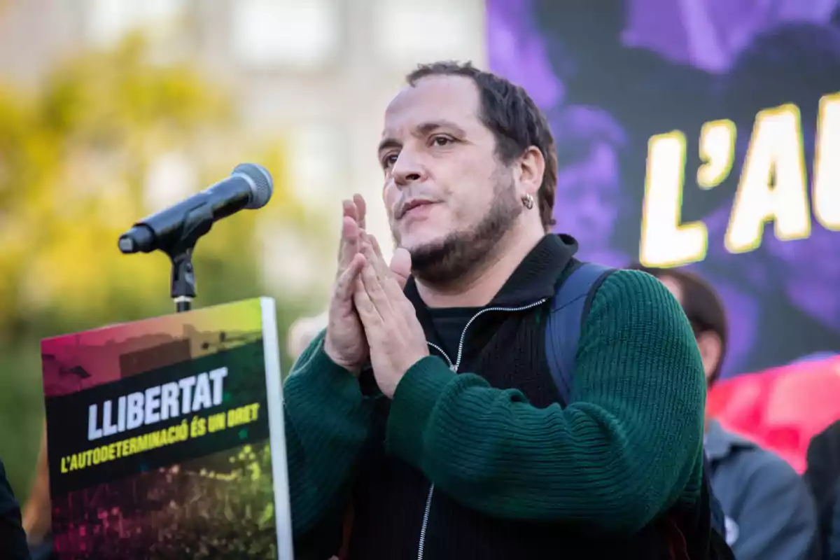 David Fernàndez parlant en un micròfon durant una manifestació amb un cartell que diu “Llibertat l'autodeterminació és un dret”.