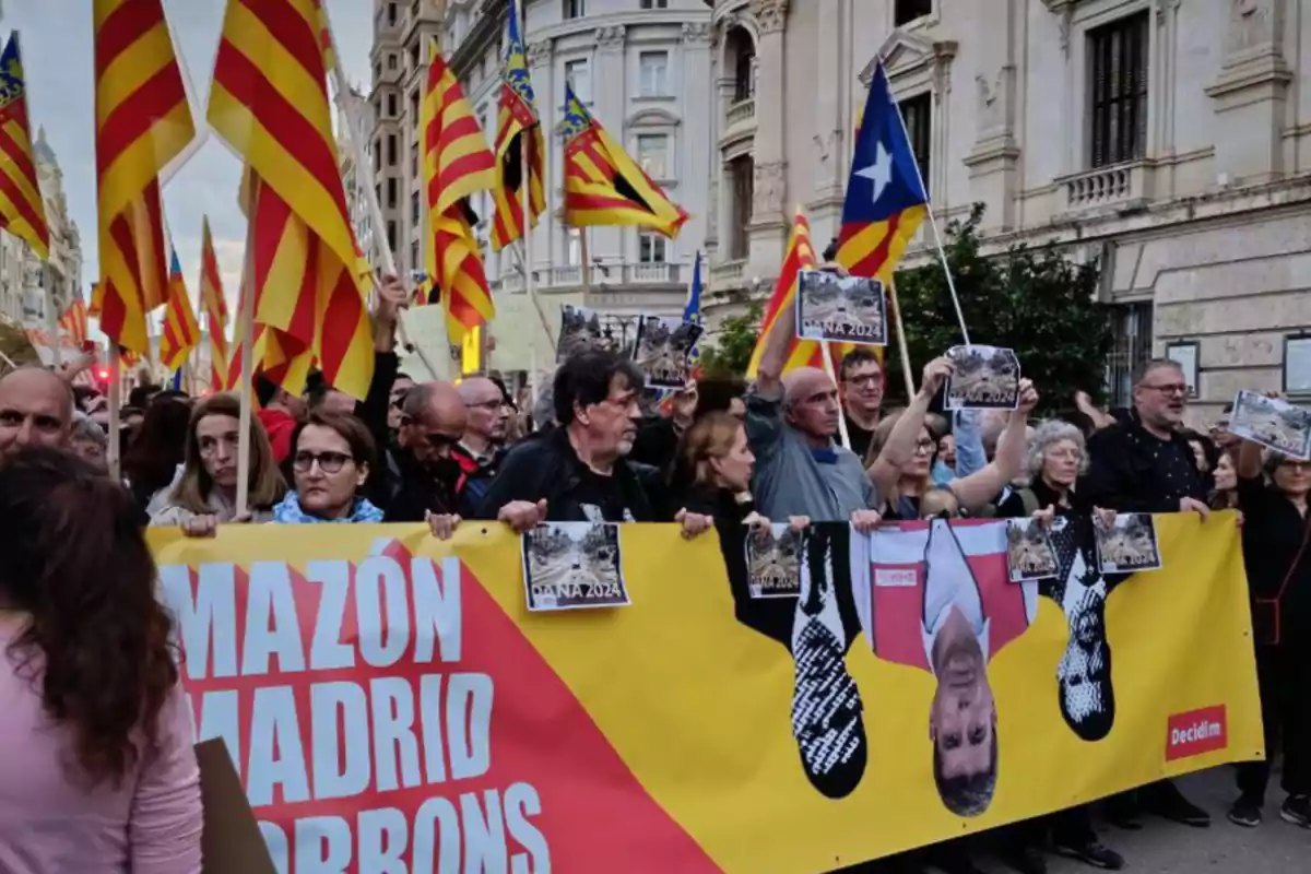 Un grup de persones participa en una manifestació al carrer, sostenint una pancarta groga i vermella amb text i diverses banderes amb franges grogues i vermelles.