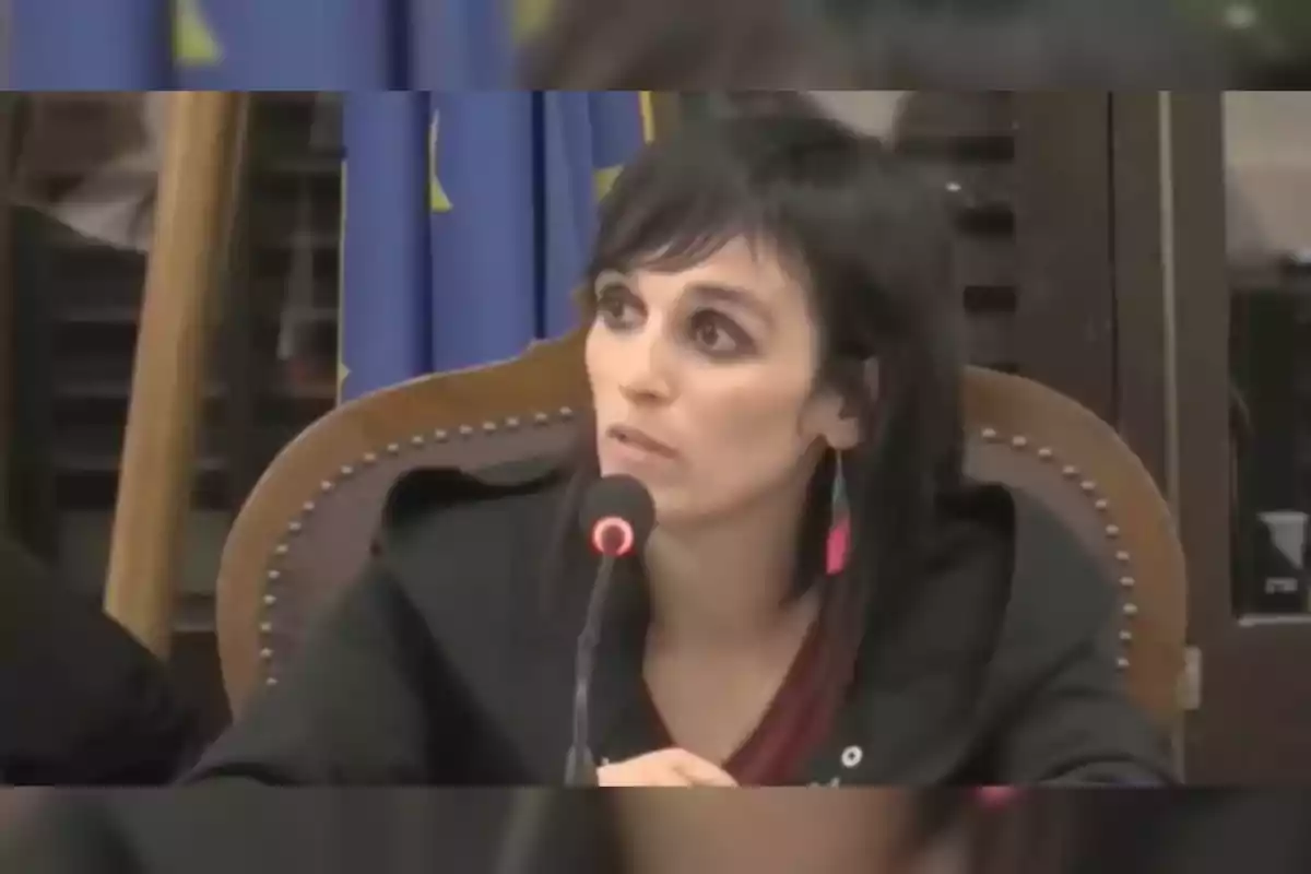 Sílvia Orriols parlant en un micròfon durant una conferència amb una bandera de la Unió Europea de fons.