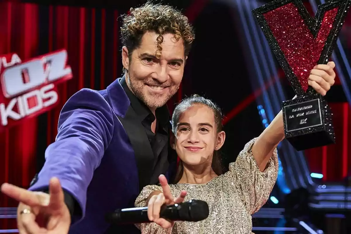 Fotografía de David Bisbal y Alira Moya sonríen y posan con un trofeo en forma de "V" en el escenario de "La Voz Kids".