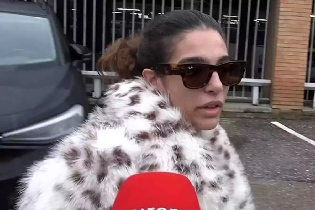 Alma Bollo wearing sunglasses and a patterned fur coat is interviewed in front of a car and a fence. Alma Bollo wearing sunglasses and a patterned fur coat is interviewed in front of a car and a fence.