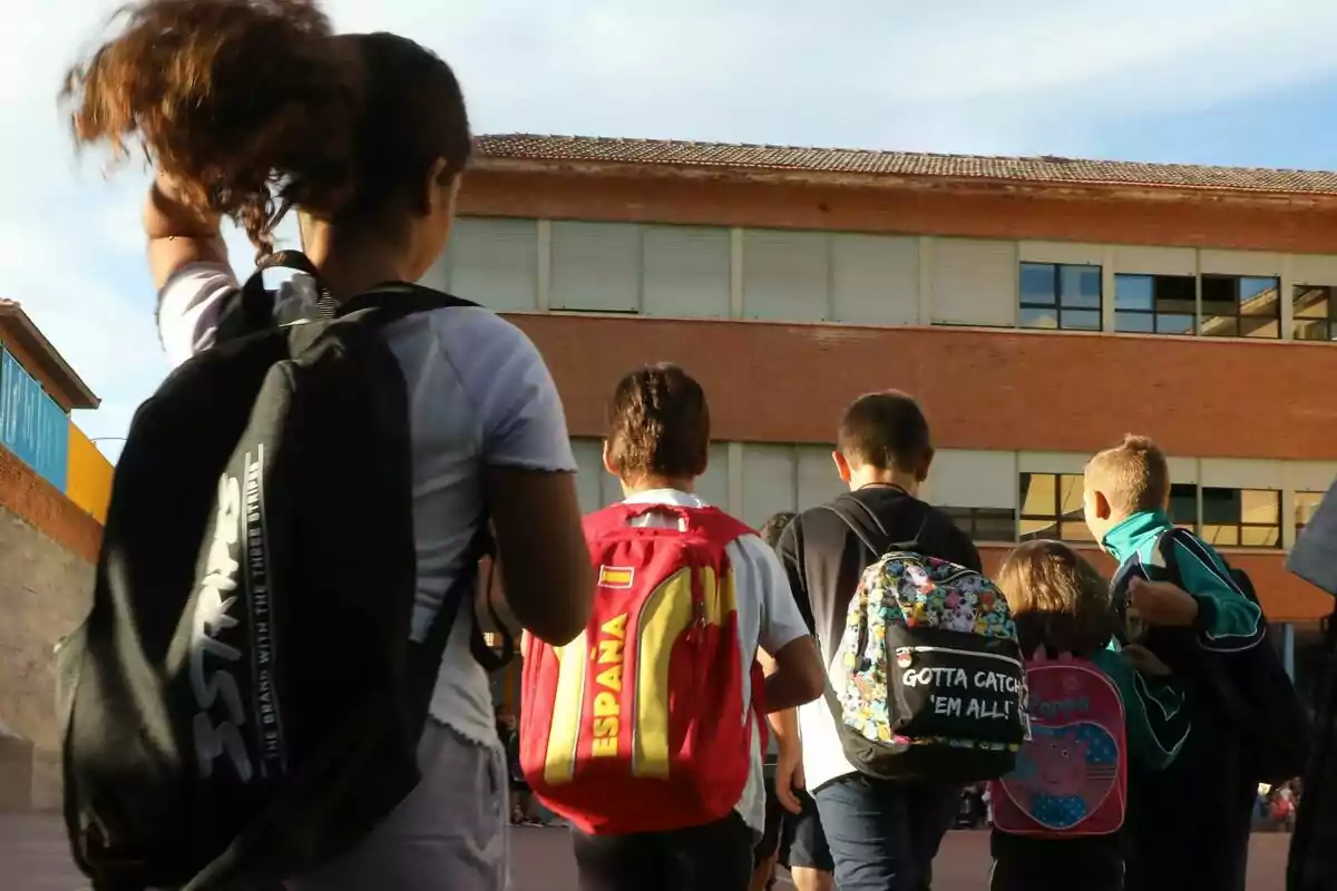 Un grup de nens amb motxilles camina cap a un edifici escolar de maó sota un cel clar. Un grup de nens amb motxilles camina cap a un edifici escolar de maó sota un cel clar.