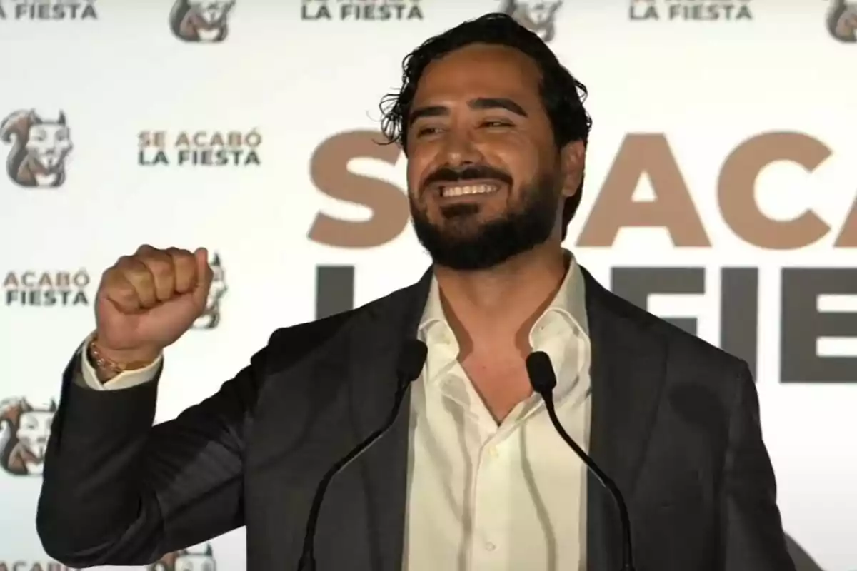 Pla mitjà d'Alvise Pérez somrient i alçant un puny amb un fons ple de logos del seu partit S'ha acabat La Festa