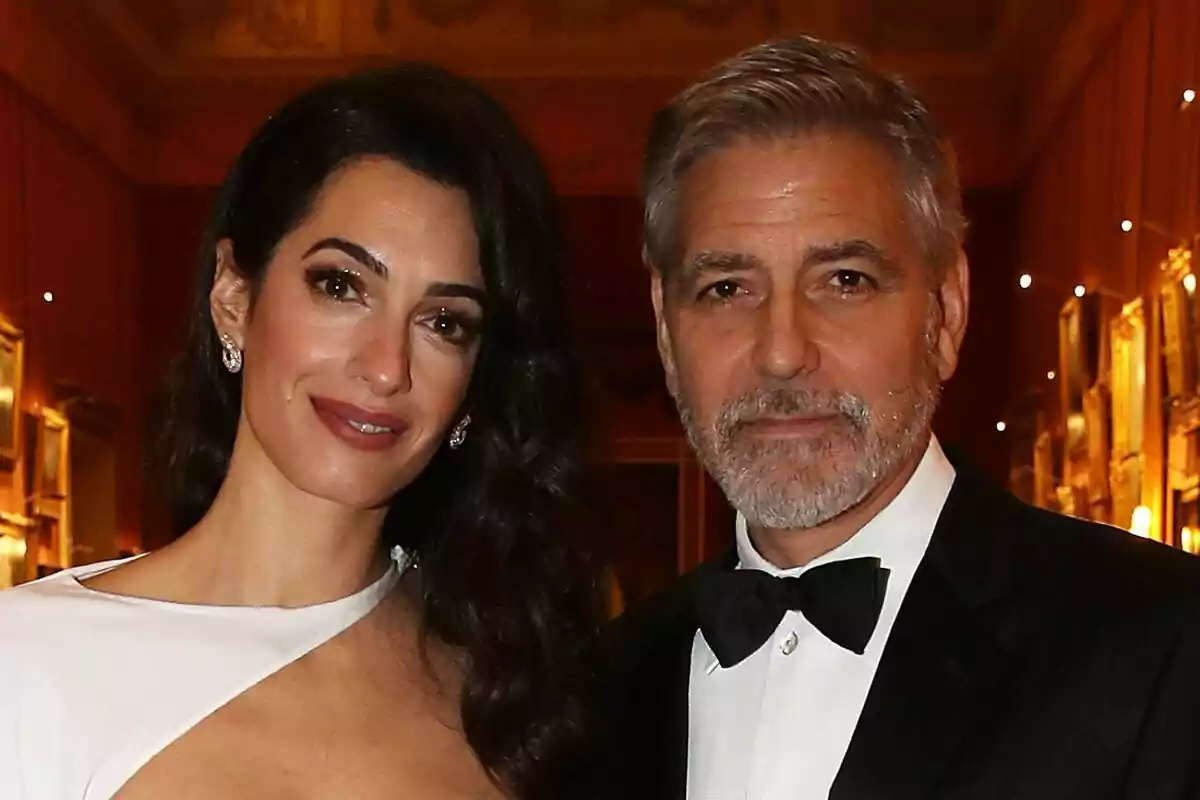 Amal and George Clooney elegantly dressed pose at a formal event with warm lighting and paintings on the walls. Amal and George Clooney elegantly dressed pose at a formal event with warm lighting and paintings on the walls.