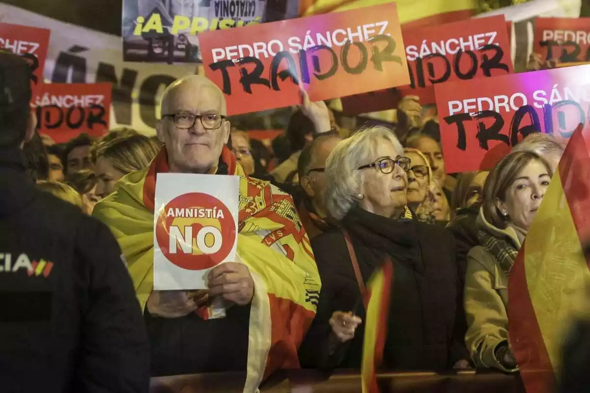 Un senyor amb una bandera d'Espanya de manera capa subjecta un cartell amb la frase 'amnistia no'. Darrere seu, diverses persones mostres cartells amb el lema 'Pedro Sánchez traïdor'