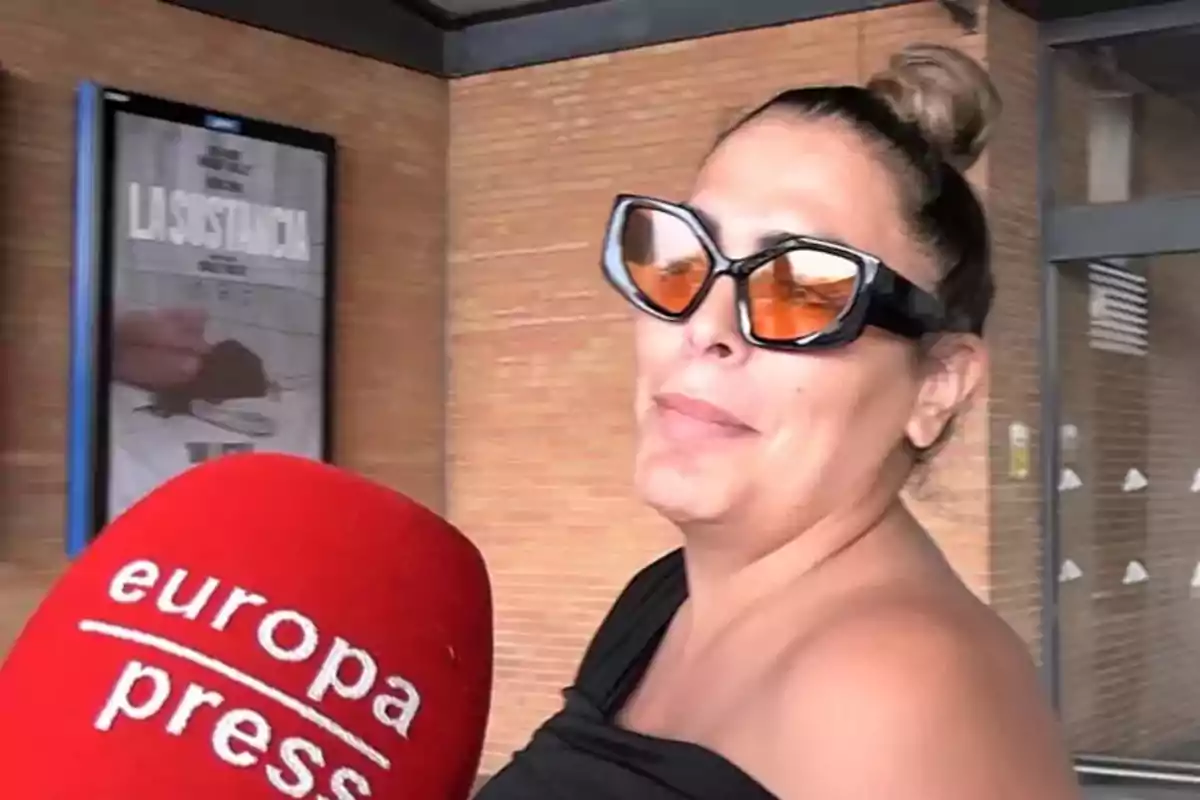 Amor Romeira con gafas de sol naranjas es entrevistada por un micrófono de Europa Press frente a un cartel de película.