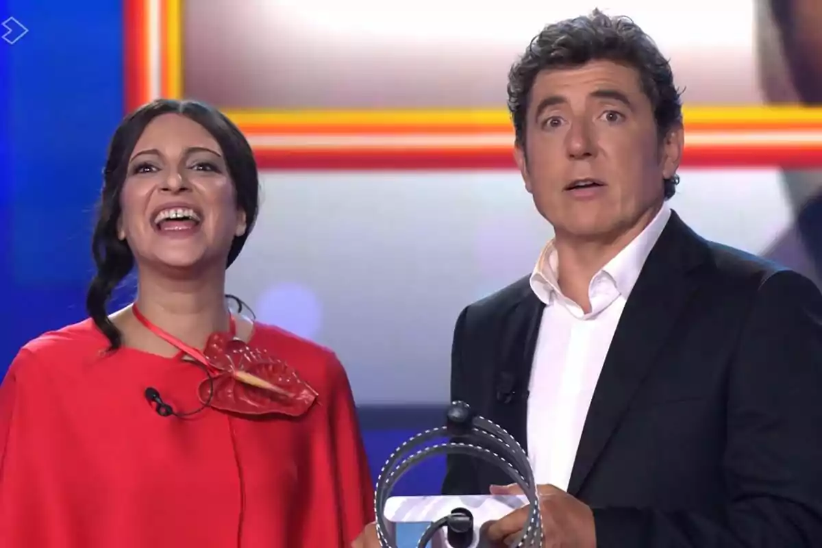 Dos presentadors a Tu Cara Me Suena, Ana Guerra somriu àmpliament i Manel Fuentes sembla sorprès, tots dos estan drets davant d’una màquina o dispositiu circular.