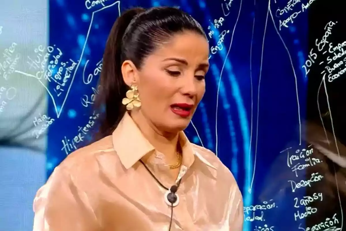 Ana Herminia con cabello oscuro recogido y aretes dorados grandes está hablando en GH DÚO con un fondo azul que tiene texto escrito en blanco.