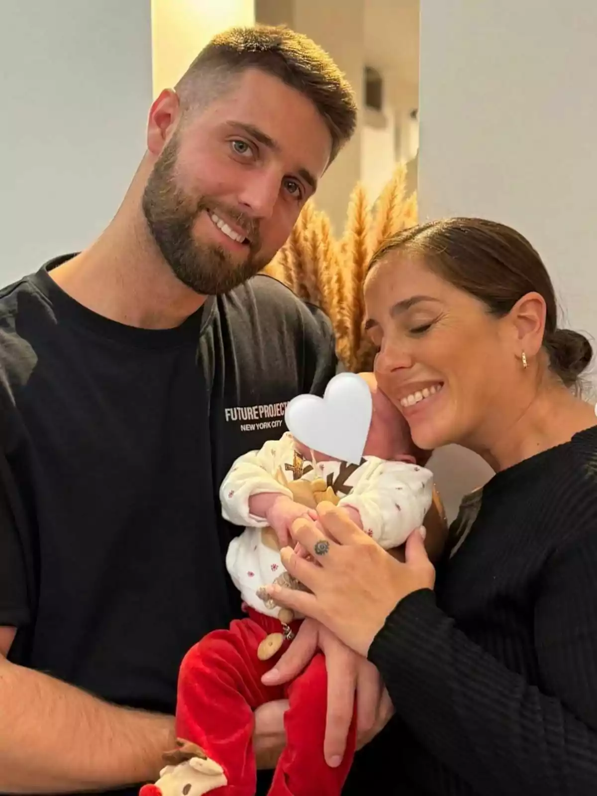 Una pareja sonriente sostiene a un bebé con un corazón blanco cubriendo su rostro.