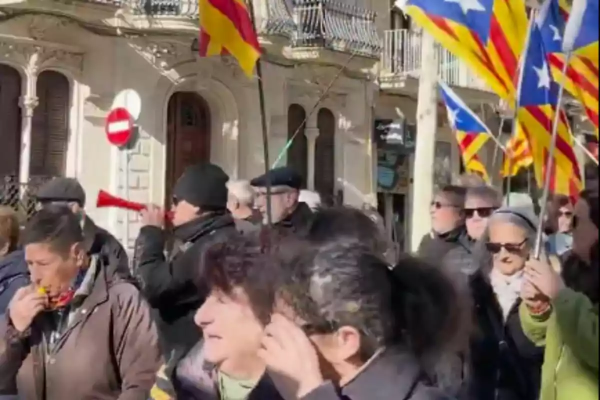 Persones participant en una manifestació amb banderes i vuvuzeles en un carrer assolellat.