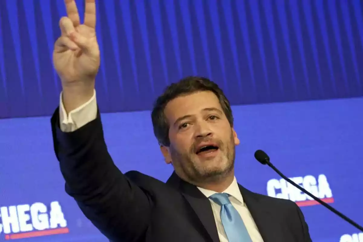 Un home amb vestit i corbata blava aixeca la mà fent un gest de victòria mentre parla davant d'un micròfon amb un fons blau que té la paraula "CHEGA".