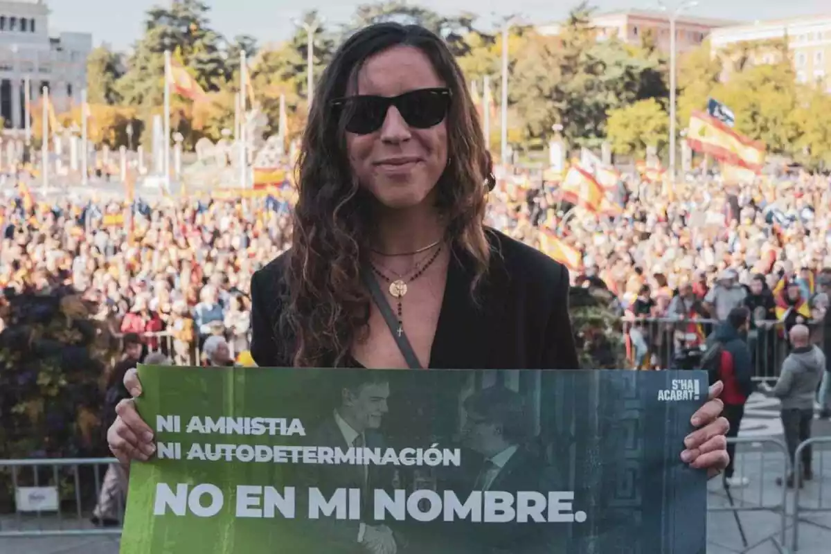Andrea Llopart somrient amb ulleres de sol i subjectant un cartell verd amb el lema 'ni amnistia ni autodeterminació, no en nom meu'. De fons, una concentració amb centenars de persones amb banderes espanyoles