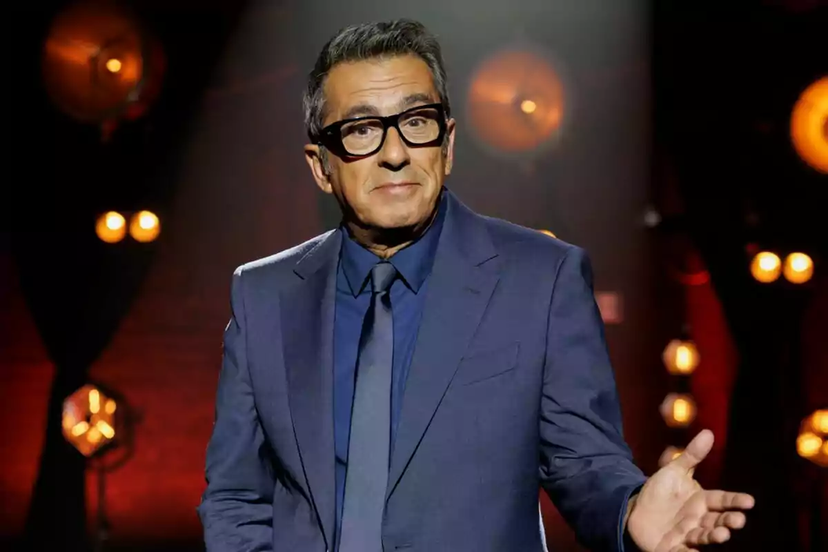 Andreu Buenafuente amb ulleres i vestit blau fosc gesticula amb una mà en un escenari il·luminat amb llums càlides a Futuro Imperfecto.