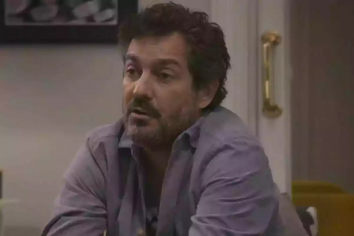 Marc Cartes with a beard and dark hair is wearing a blue shirt and is sitting in a room with a door in the background.