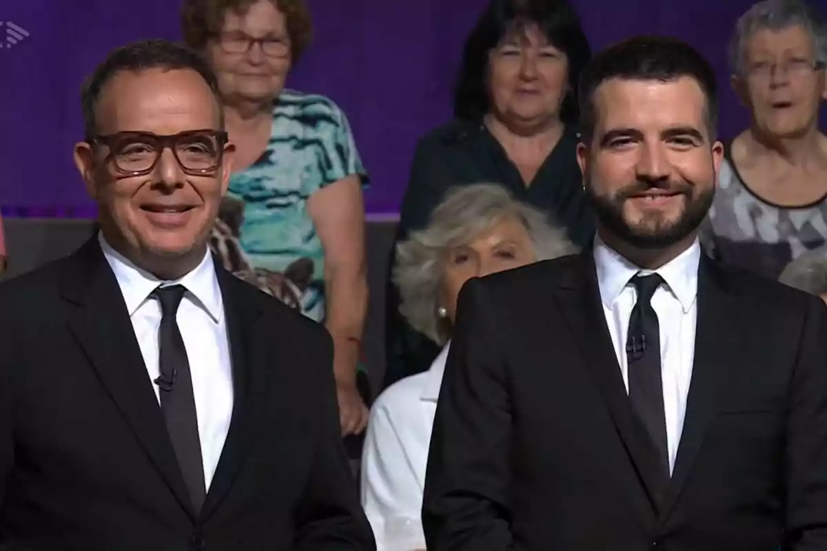 Àngel Llàcer i Ricard Ustrell vestits amb vestit i corbata negra somriuen davant d'una audiència en un estudi de televisió de Col·lapse. Àngel Llàcer i Ricard Ustrell vestits amb vestit i corbata negra somriuen davant d'una audiència en un estudi de televisió de Col·lapse.