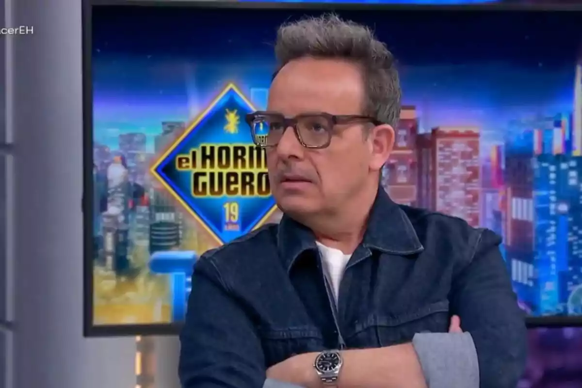 Ángel Llàser con gafas y chaqueta de mezclilla está en un set de televisión con un fondo que muestra el logo de "El Hormiguero".