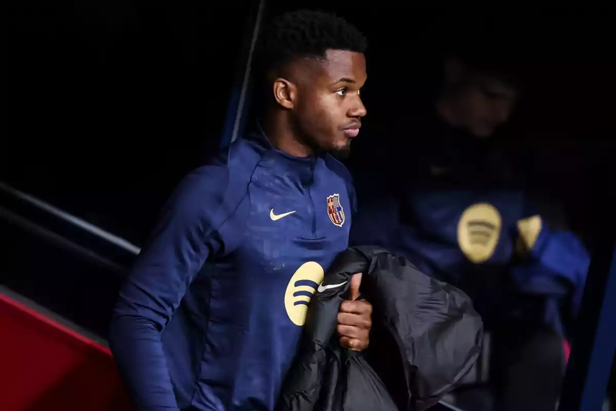 A player in a navy blue FC Barcelona uniform holds a black jacket while standing in a dark area.