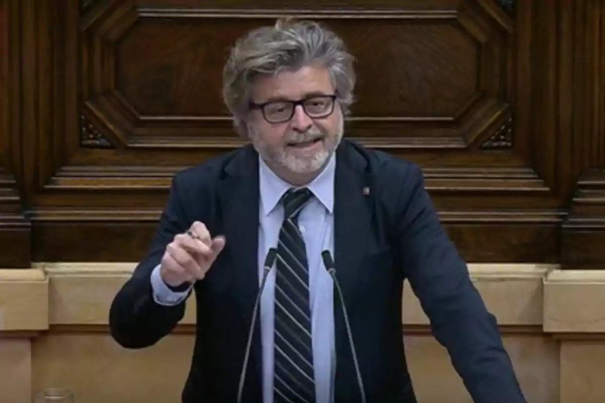 Un hombre con barba y gafas habla en un podio con un fondo de madera.