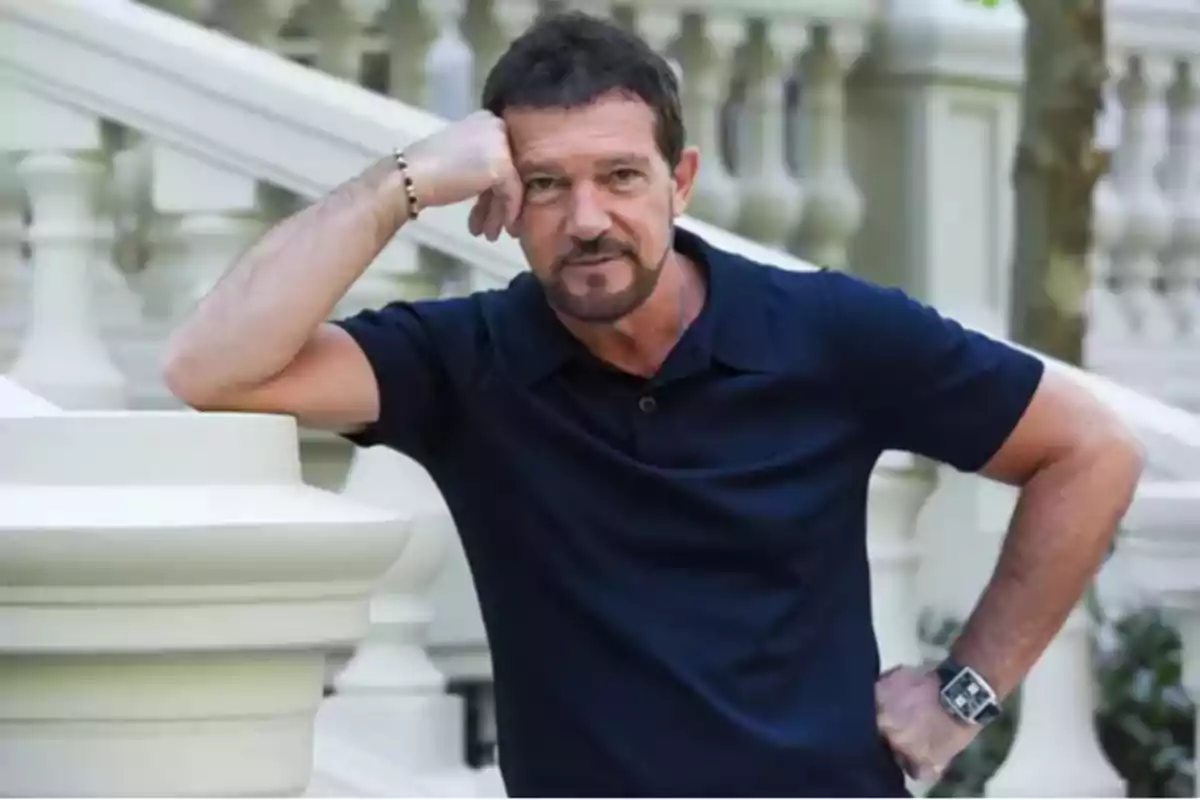 Antonio Banderas con barba y bigote, vestido con una camiseta azul marino, apoyado en una barandilla blanca.